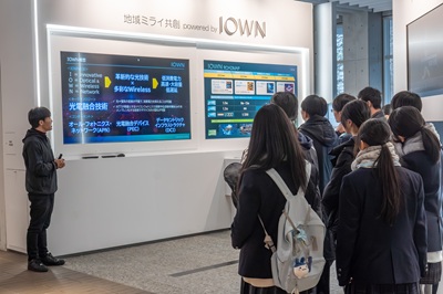 A scene of touring NTT e-City Labo. At the top of the screen, it says "Regional Future Co-creation IOWN," with an employee explaining on the left and participants receiving the explanation on the right.