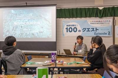 Cena da primeira palestra da Conferência Jovem pelo Clima. À direita da tela onde os materiais foram projetados está o palestrante Jun Hasegawa, e na frente dele os participantes estão sentados em cadeiras ouvindo a palestra.